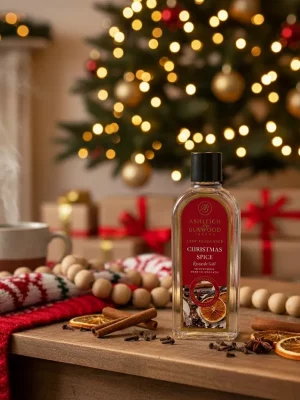 Butelka A&B_Christmas Spice stoi na drewnianym stole z suszonymi plastrami pomarańczy i laskami cynamonu, przed udekorowaną choinką i zapakowanymi prezentami, z parującym kubkiem i przytulnym dzianinowym kocem w pobliżu.