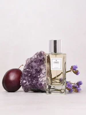 Flakon perfum nastroje_bliskosc_i_czulosc_2 ze srebrną nakrętką jest wyświetlany obok kryształów ametystu, ciemnej śliwki i małych fioletowych kwiatów na jasnej powierzchni z gładkim tłem.