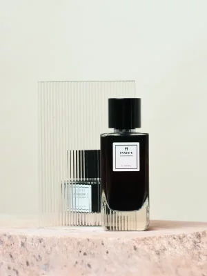 Perfumy nastroje_niezaleznosc_i_indywidualizm_2 to elegancki czarny flakon z prostokątną etykietą i czarną nakrętką, umieszczony na teksturowanej powierzchni. Prążkowany, półprzezroczysty szklany panel odbija jego kształt na miękkim, neutralnym tle.