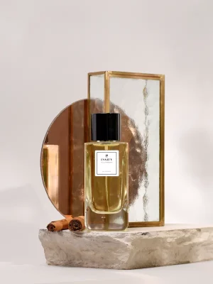 Butelka perfum z napisem „nastroje_nostalgia_i_wspomnienia_2” stoi na kamiennej płycie obok dwóch lasek cynamonu, a w tle widać okrągłe lustro z brązu i panel ze szkła o fakturze. Scena jest minimalistyczna i elegancka.